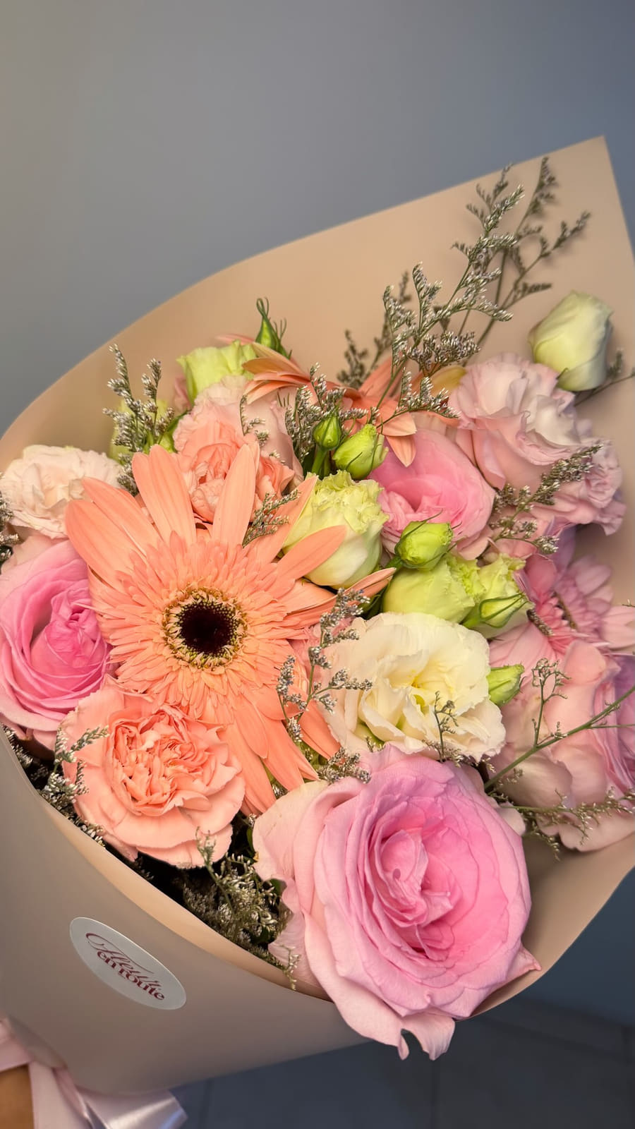 A close-up of a Fleur en Route bouquet: peach gerbera, pink China roses, lisianthus and misty in blush wrap with the brand sticker